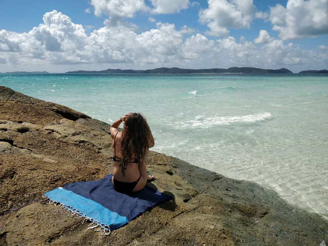 A person sitting on a rocky shoreline, using a blue mystics beach towel by the clear water and sky.