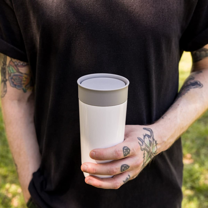 A stainless steel coffee cup in white with a grey lid, held by a person against a green background.
