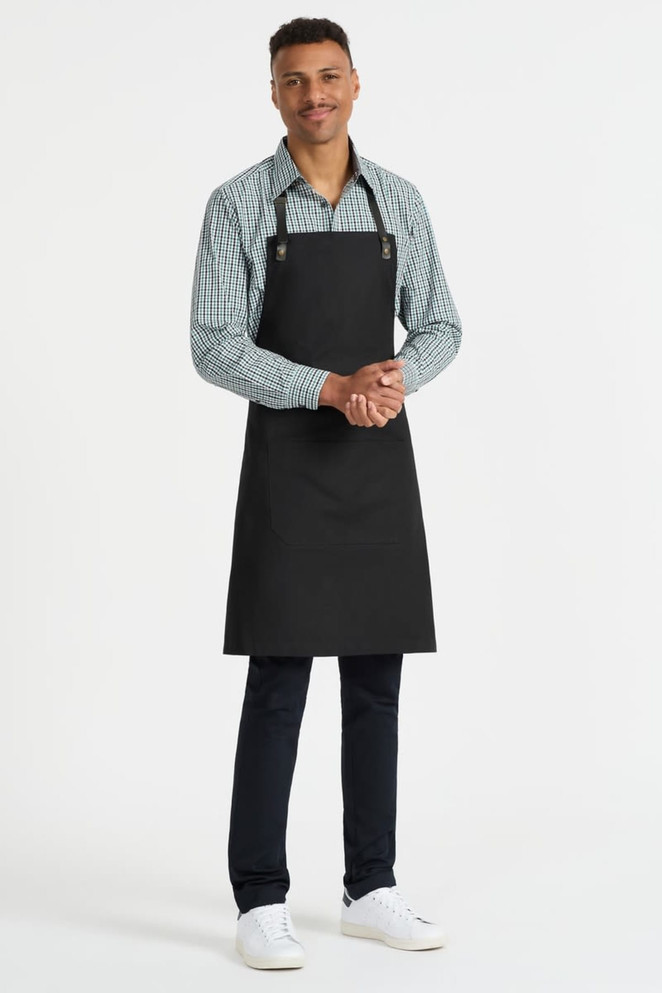 A man wearing a black apron over a green and white checkered button-up shirt, standing in a neutral background.