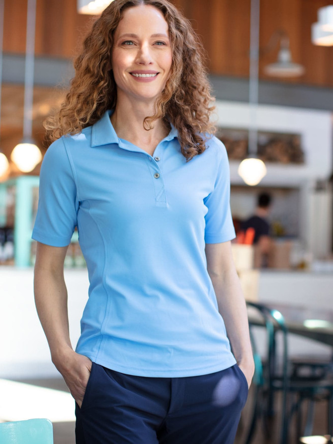 A woman stands in a café wearing a light blue polo shirt paired with dark trousers. The shirt features a logo.