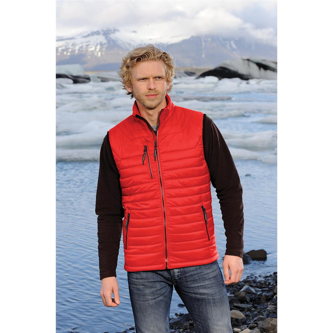 A man wearing a red thermal vest over a black long-sleeve shirt, standing by a glacial landscape.