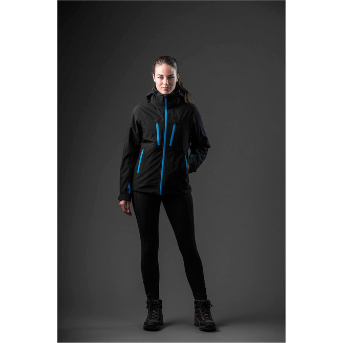 A woman stands in a black jacket with blue accents and black leggings, featuring a logo. The background is dark.