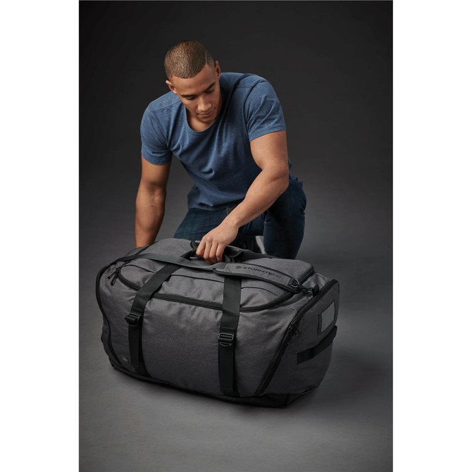 A man kneels beside a large grey duffle bag with black straps, featuring a logo on the front.