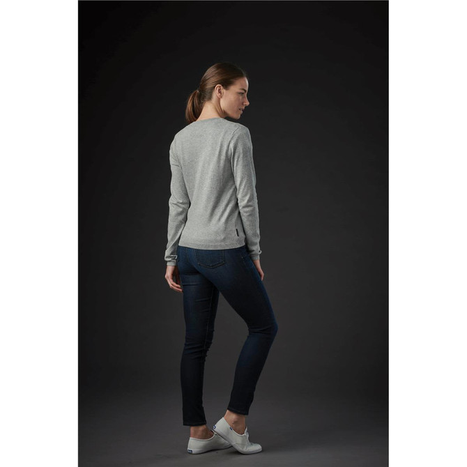 A woman wearing a grey cardigan and dark jeans, standing with her back to the camera on a dark background.