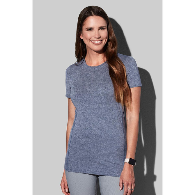 A woman wearing a blue recycled sports T-shirt, smiling and posing against a grey background. The shirt features a logo.