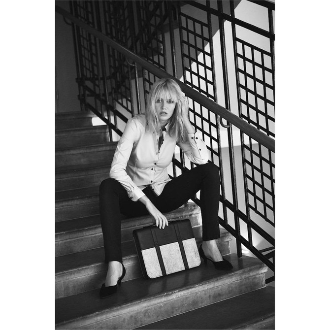 A model wearing a white button-up shirt sits on stairs, holding a stylish black and beige bag. Black pants and shoes complete the look. A model wearing a white button-up shirt sits on stairs, holding a stylish black and beige bag. Black pants and shoes complete the look.