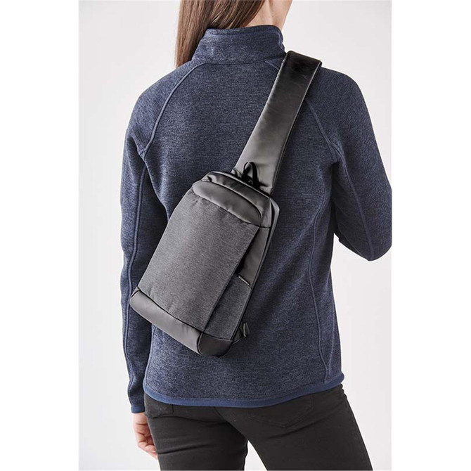 A grey and black sling backpack worn over the shoulder, displayed on a person in a blue jacket.