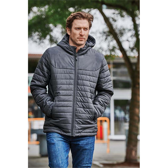 A man wearing a dark grey quilted hoody, walking outdoors surrounded by greenery. The hoody features a logo.