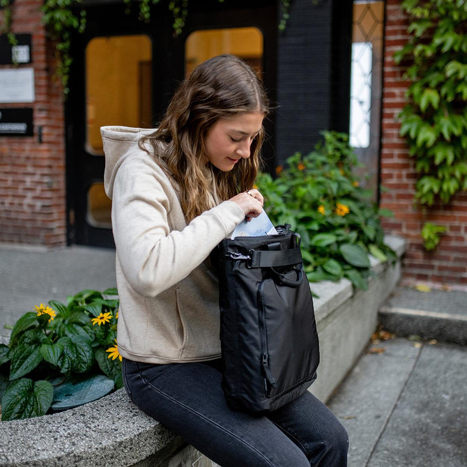 A woman interacting with a black Meridan Tote Backpack while sitting on a stone ledge with greenery around.