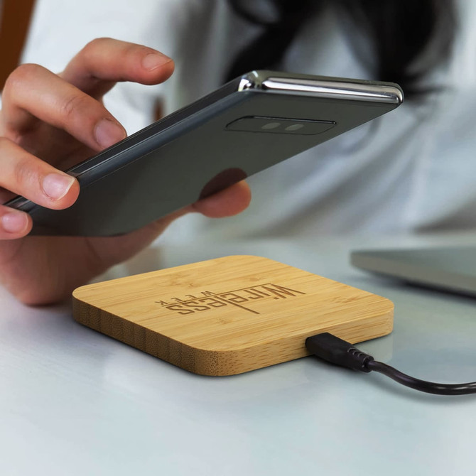 A bamboo wireless fast charging hub with a connected cable, placed on a desk, alongside a person holding a smartphone.