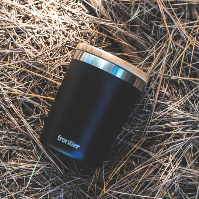 A drink bottle in matte black with a wooden lid, featuring a simple logo on the side, resting on straw.