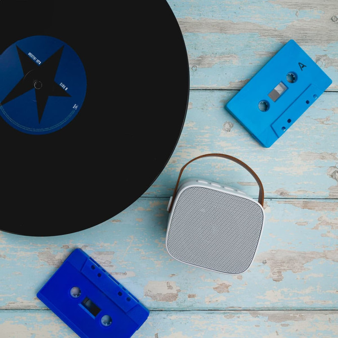 A white mini speaker with a brown handle, accompanied by blue cassette tapes and a black vinyl record on a wooden surface.