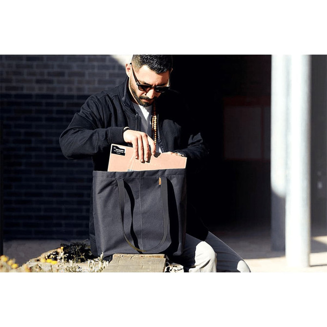 A black tote bag with long handles is being carried by a person. The bag contains a notebook and has a logo.