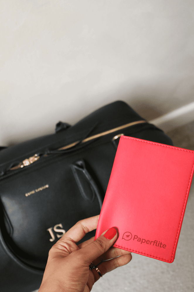 A bright pink vegan leather passport wallet is held in hand, with a black duffel bag in the background.