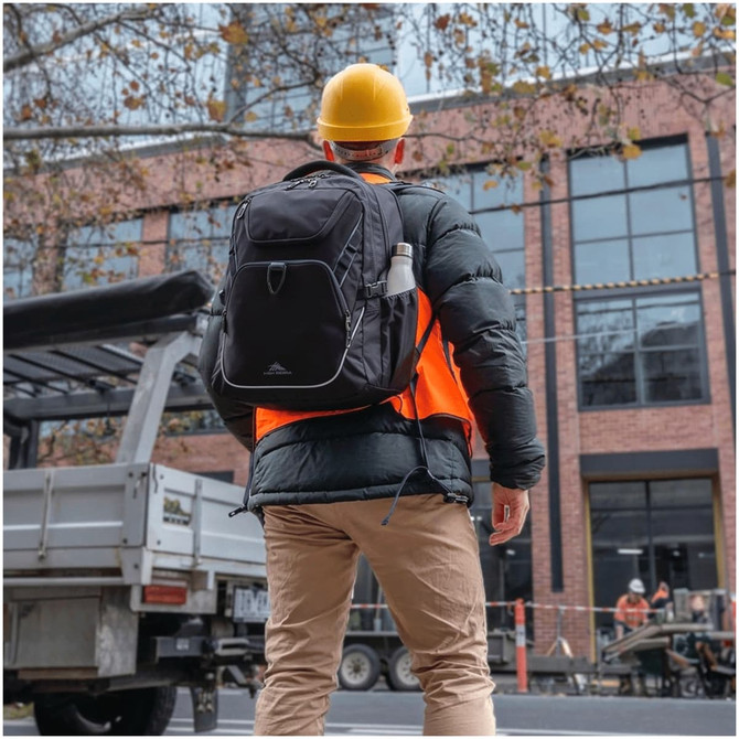A black High Sierra Access 4.0 backpack rests on the back of a person wearing a hard hat and bright orange vest.