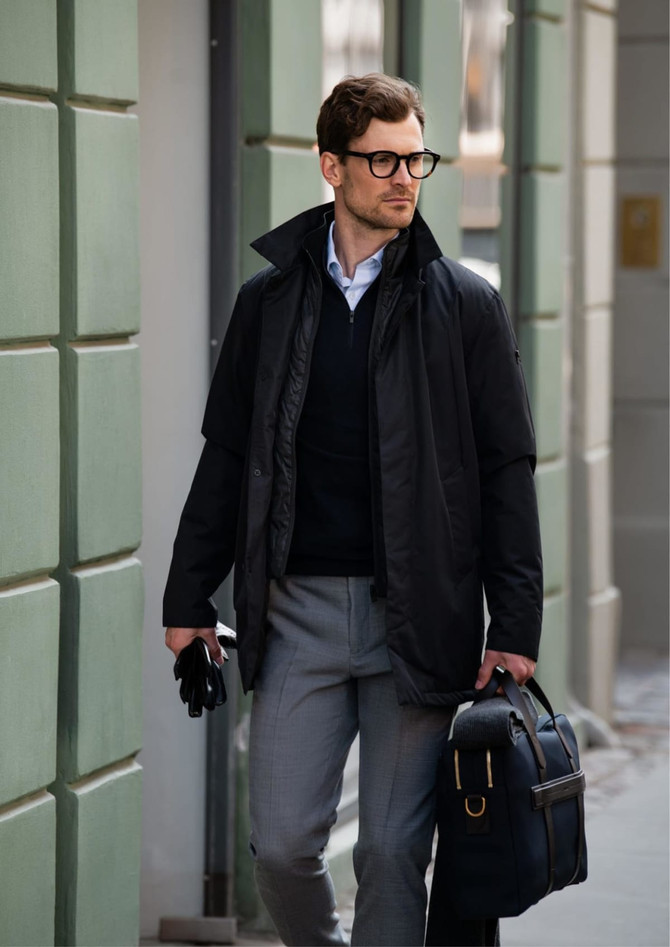A black jacket worn over a dark sweater, paired with grey trousers and a stylish bag, featuring a logo.