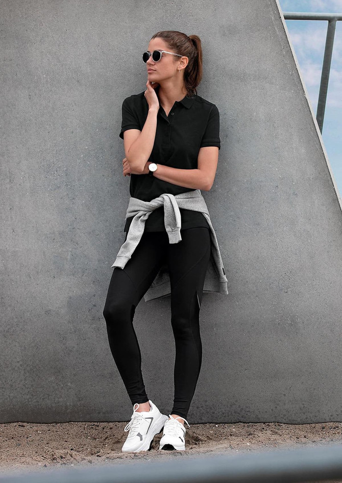 A model wearing a black polo shirt with a grey sweater tied around her waist, paired with black leggings and white sneakers.