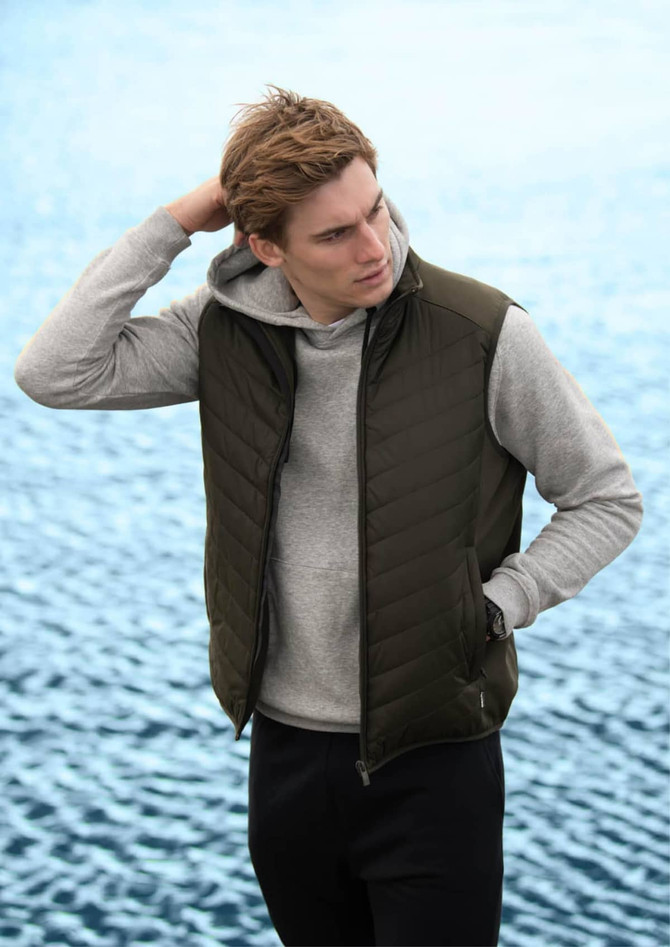 A green puffer vest worn over a grey hoodie, modelled by a young man against a water background.