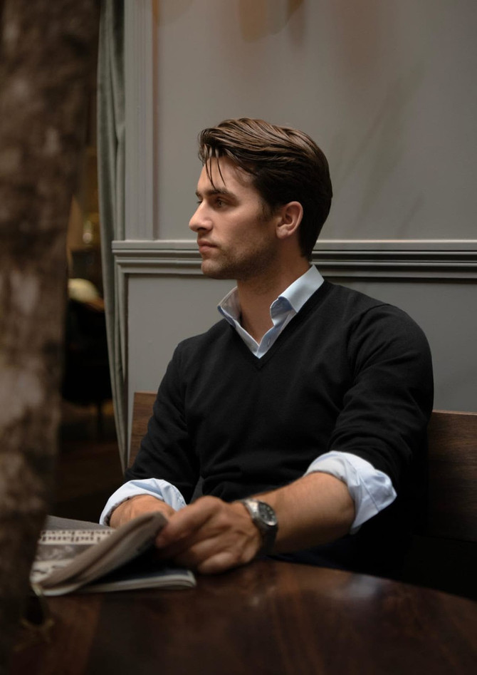 A man wearing a black V-neck jumper sits at a wooden table, holding a newspaper.