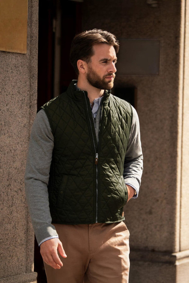 A man wearing a dark green quilted vest over a grey long-sleeve shirt, standing outside a building.