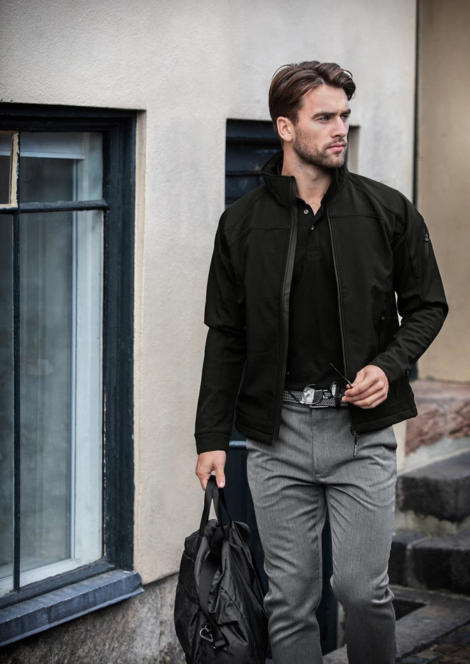 A man wearing a black soft shell jacket, carrying a bag, stands near a building with windows.