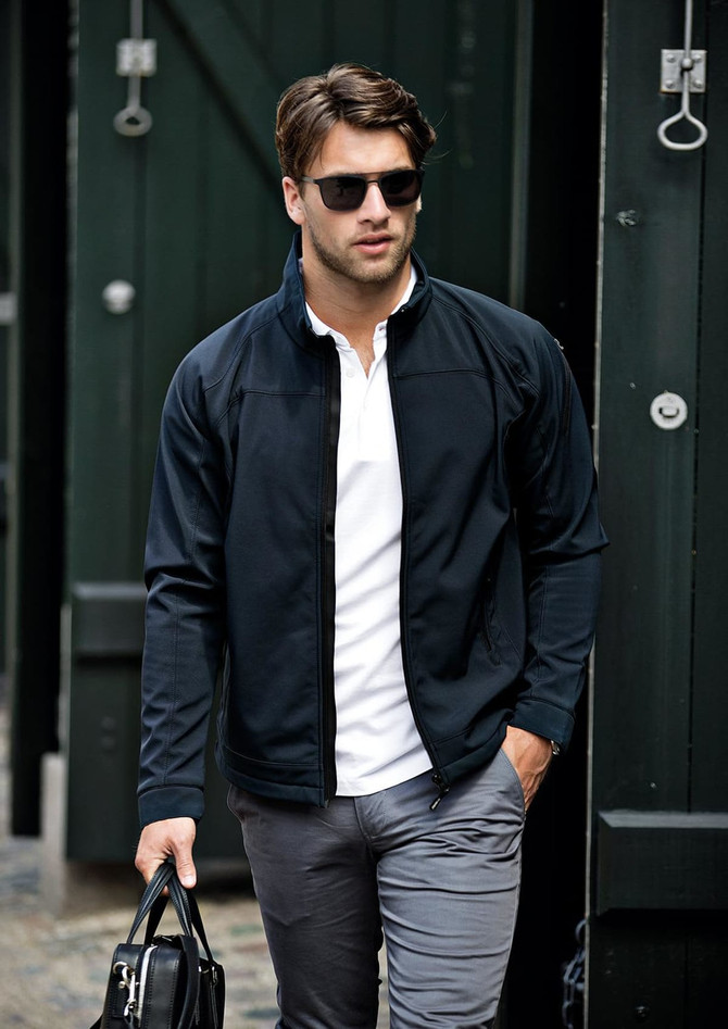 A man in a black soft shell jacket walks confidently, wearing sunglasses and holding a bag.