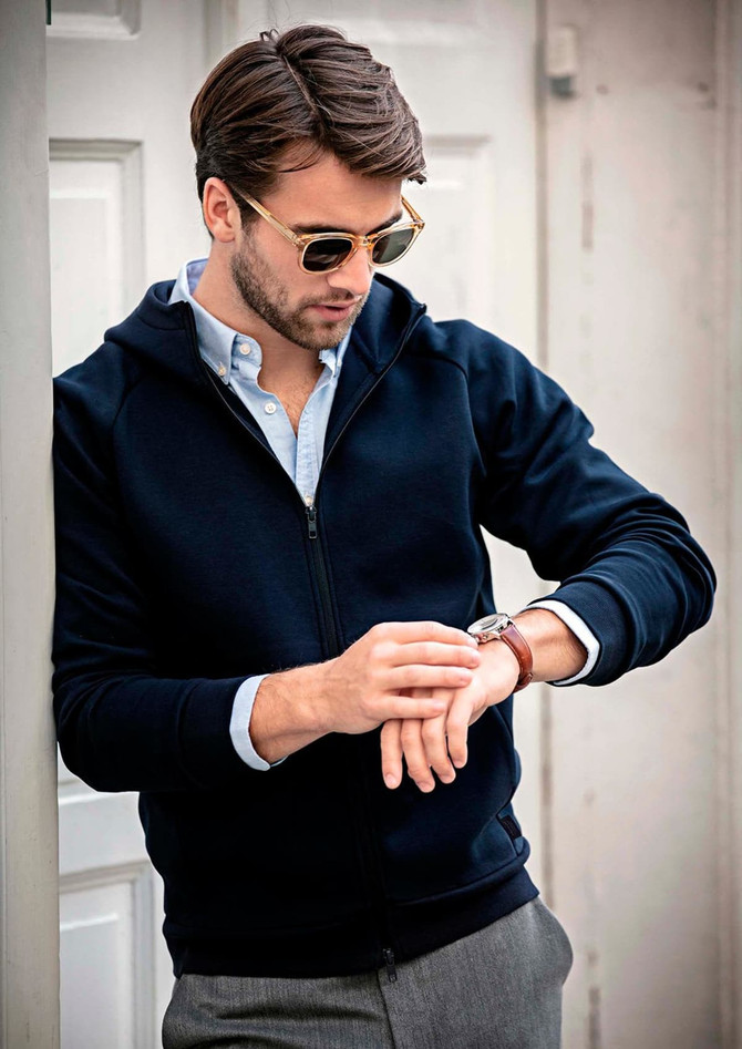 A navy hoodie jacket with a zip, featuring a hood, worn by a man checking his watch. It has a logo.