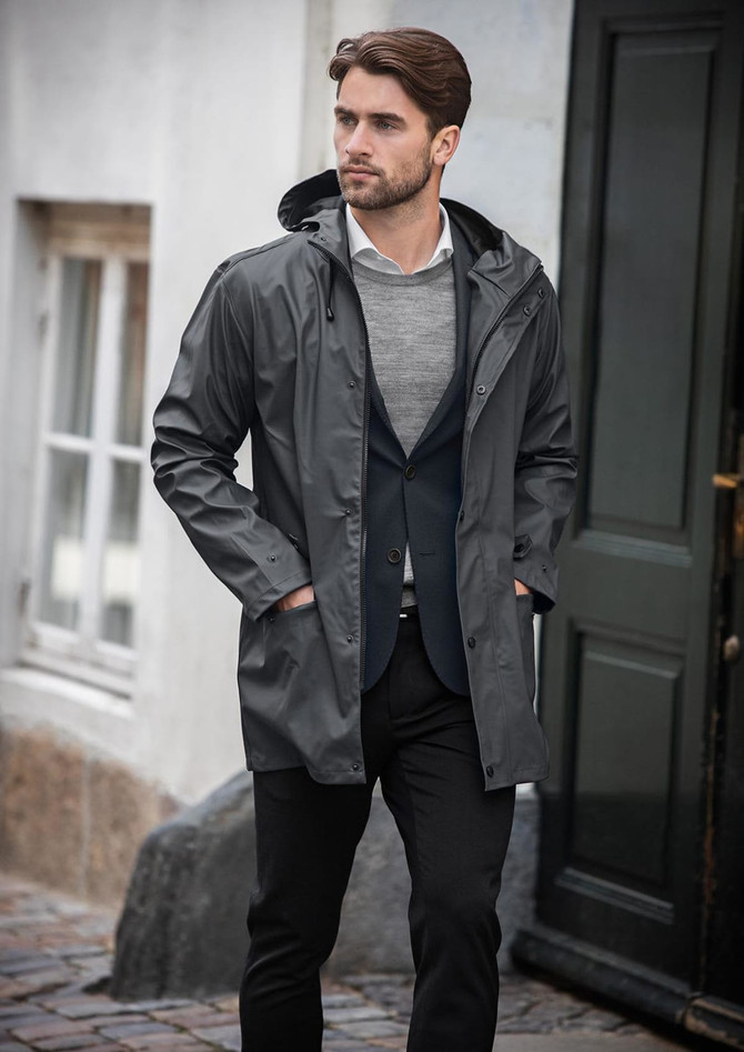 A grey rain jacket with a hood, worn by a man in smart casual attire, standing near a door.