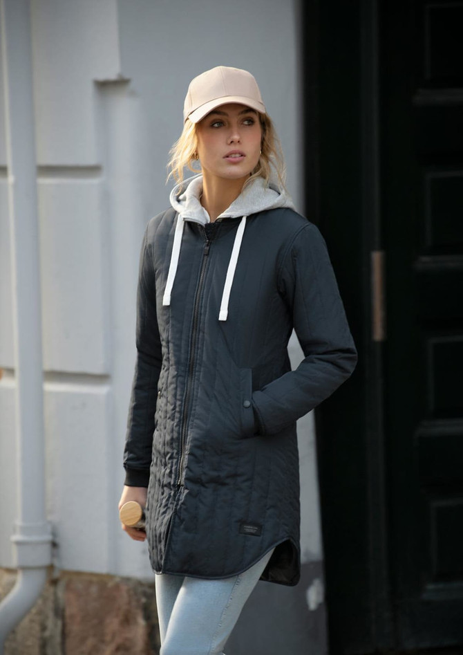 A woman wearing a grey puffer jacket with a hoodie underneath, paired with a beige cap and jeans. The jacket features a logo.