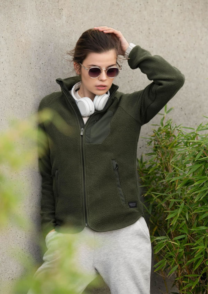 A green women's jacket with a zip front and pockets, worn over a grey hoodie, paired with sunglasses and headphones.