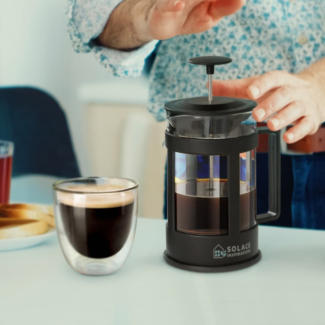 A small black coffee plunger sits on a table next to a glass of brewed coffee. The background features a blurred hand.