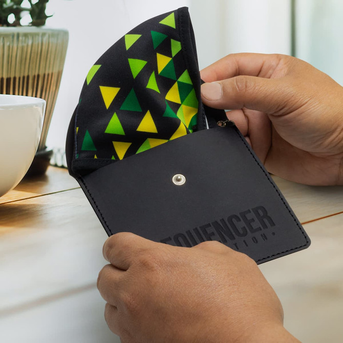 A black key ring pouch with a colourful triangular pattern inside, held in a person's hands near a plant and cup.