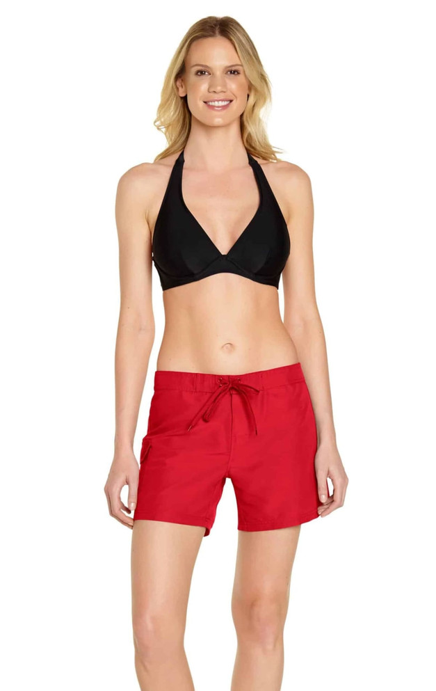 A woman models red cargo board shorts, paired with a black halter bikini top. The shorts feature a drawstring waist and a logo.