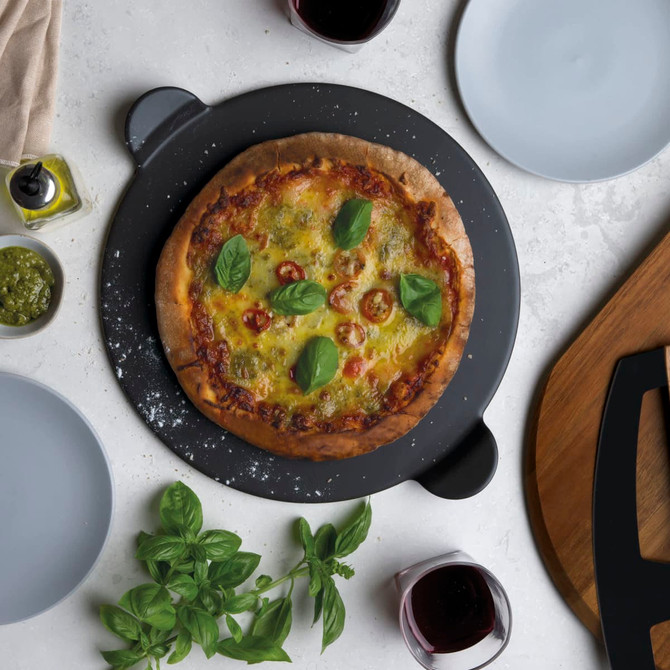 A pizza on a black serving stone, garnished with basil and cherry tomatoes, surrounded by plates and wine glasses. A pizza on a black serving stone, garnished with basil and cherry tomatoes, surrounded by plates and wine glasses.