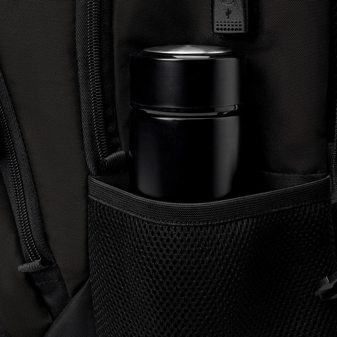A black thermos is nestled in a mesh pocket of a black backpack featuring various zippers and compartments.