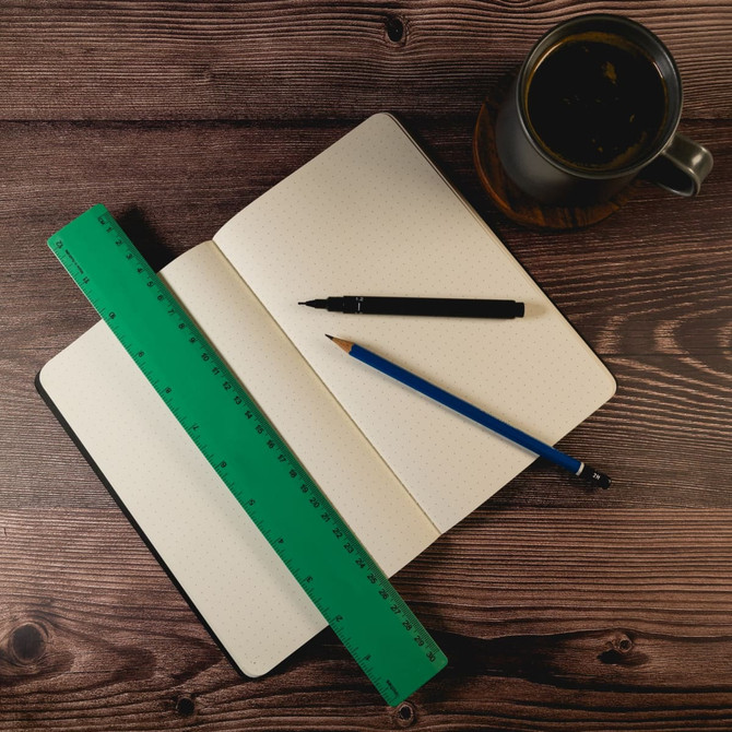 A green 30cm recycled plastic ruler rests beside an open dotted notebook, a black pen, and a blue pencil, with a coffee cup nearby. A green 30cm recycled plastic ruler rests beside an open dotted notebook, a black pen, and a blue pencil, with a coffee cup nearby.