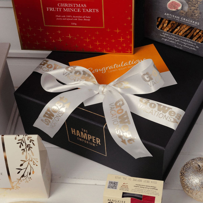 A black gift box tied with a white ribbon and a card, surrounded by festive food items and packaging.