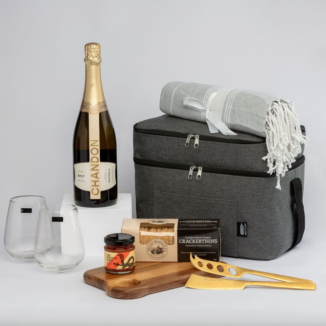 A grey picnic cooler bag with a bottle of sparkling wine, glasses, a throw blanket, gourmet snacks, and a cheese knife.