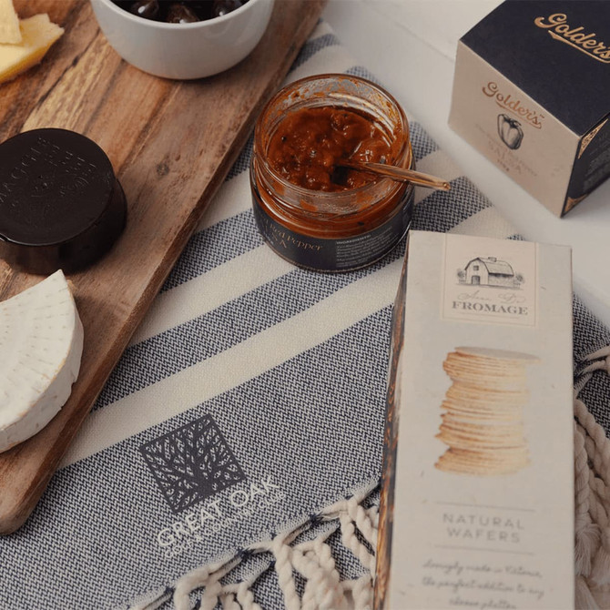 A wooden board with cheese, a jar of spread, and a box of wafers on a textured cloth with a logo.