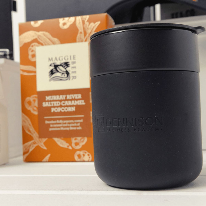 A black insulated drink container with a lid beside a box of salted caramel popcorn, featuring a logo on the container.