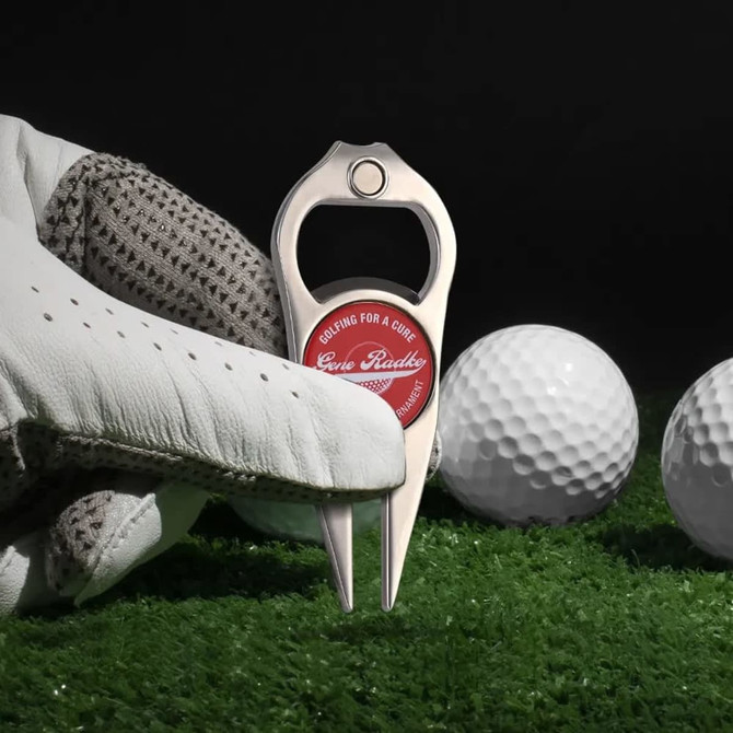 A silver divot tool with a bottle opener and a red logo, placed beside three golf balls on green grass.