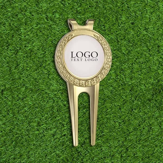 A gold golf divot tool with a circular ball marker, resting on green grass. The marker features a logo.