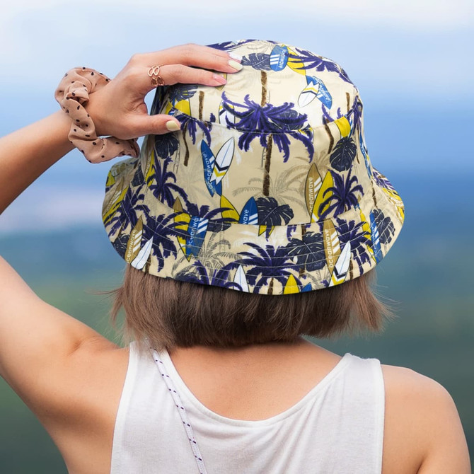 A bucket hat with a tropical print in beige and blue, featuring palm trees and surfboards. It has a logo on the side.