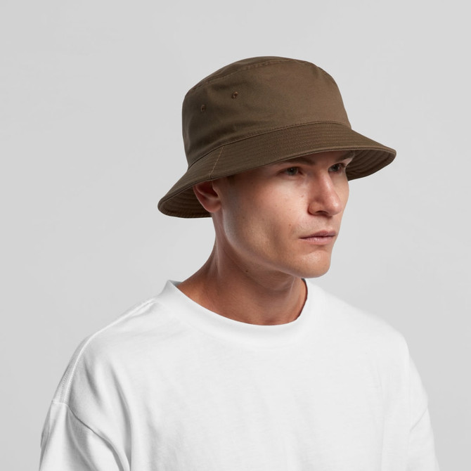 A brown bucket hat with a wide brim, displayed on a model wearing a white shirt. Features a logo on the hat.