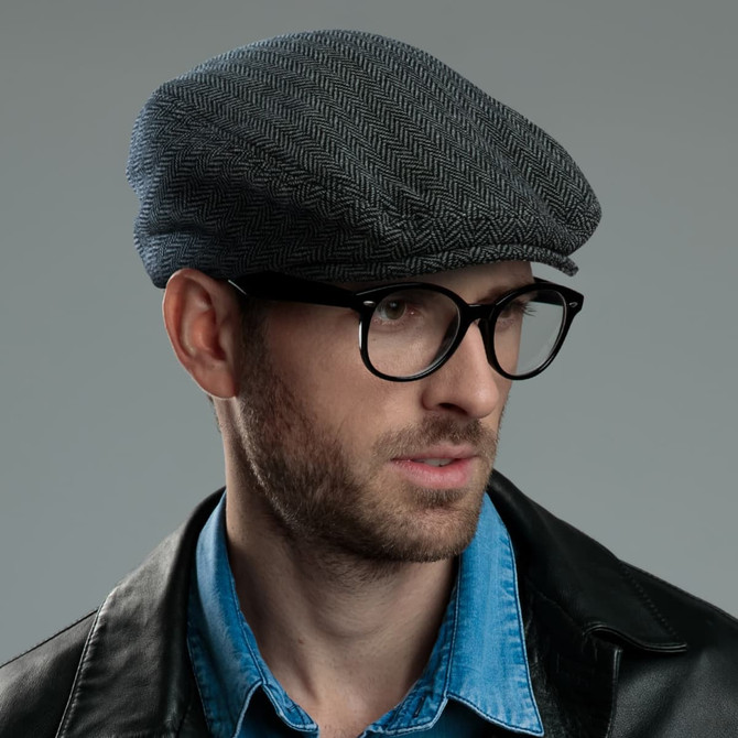 A flat cap in a herringbone pattern, predominantly grey, worn by a model with glasses. Features a logo.