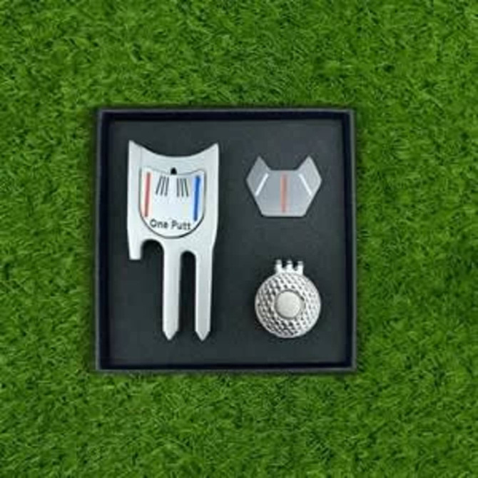 A magnetic cap clip, ball marker, and divot tool set displayed in a black box on grass. The products are silver with coloured accents.
