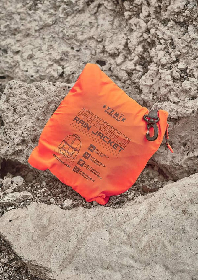 An orange packable rain jacket neatly folded and resting on rocky terrain, featuring a logo and product details.