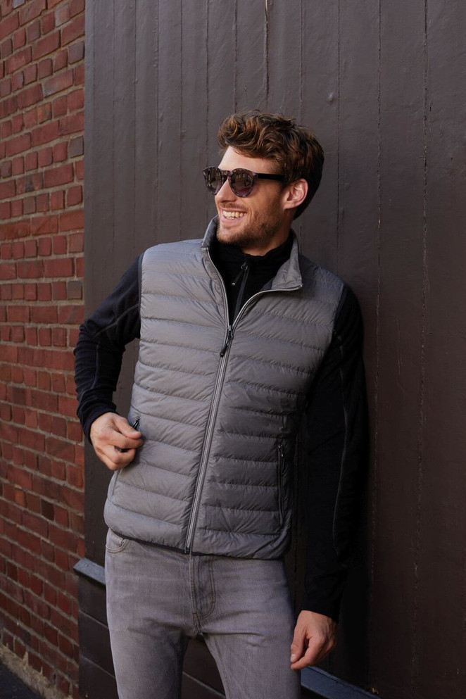 A man wearing a grey down vest stands against a brick wall, smiling and wearing sunglasses. The vest has a logo.