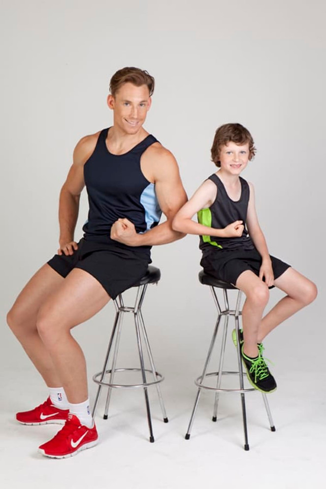 A navy and green singlet worn by an adult and a child, both seated on stools, showcasing their muscles.
