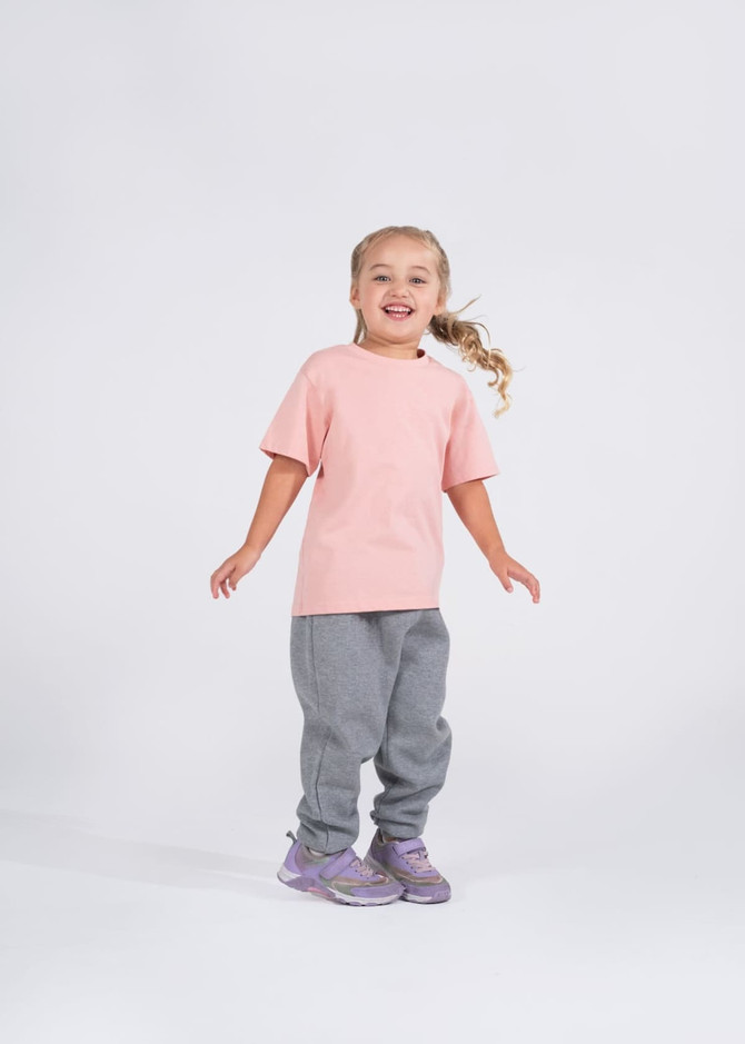 A smiling girl in a light pink T-shirt and grey sweatpants stands on a white background, with a logo visible on the shirt.